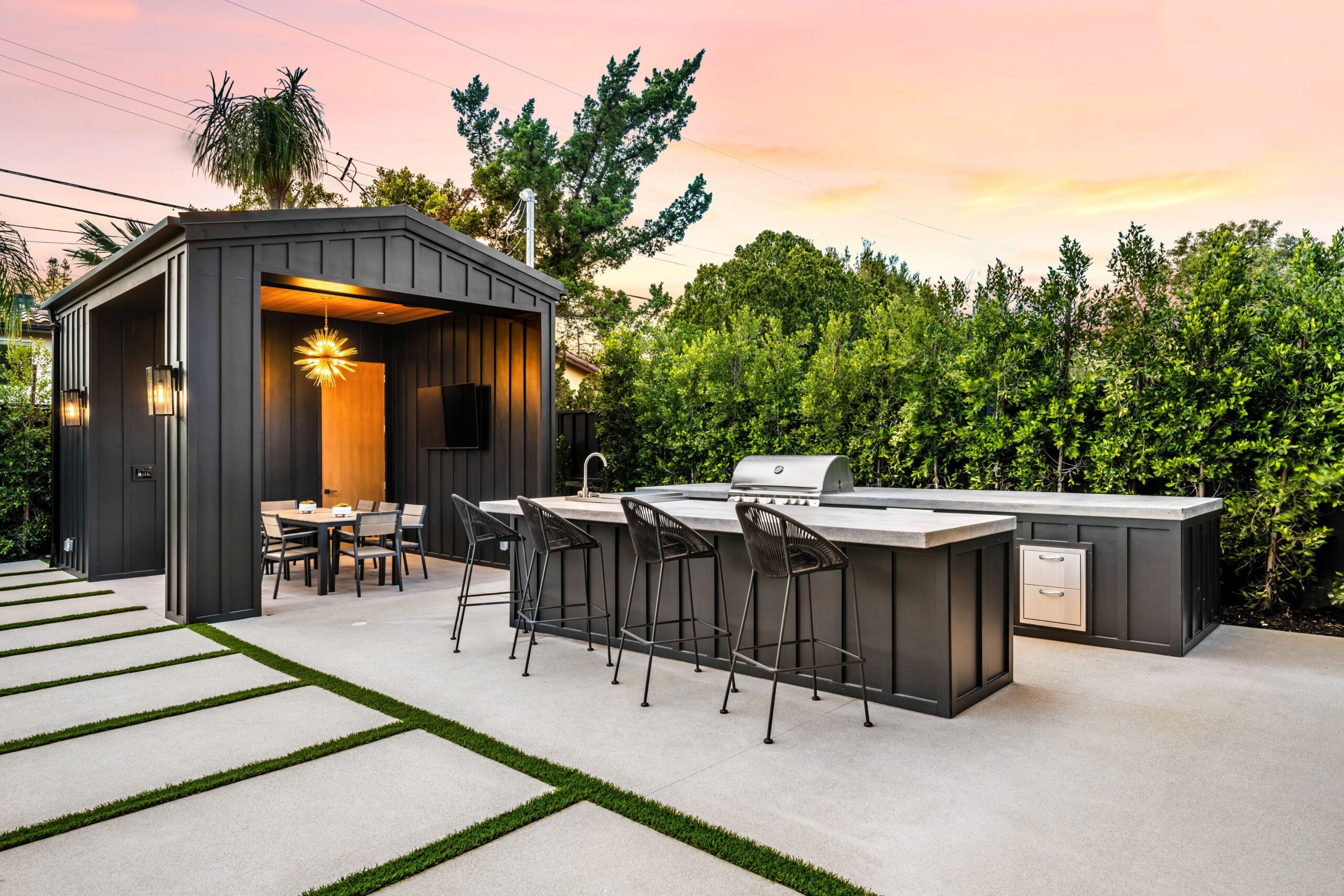 Modern outdoor kitchen and dining area with a covered table, bar seating, built-in grill, and lush green hedge at sunset.