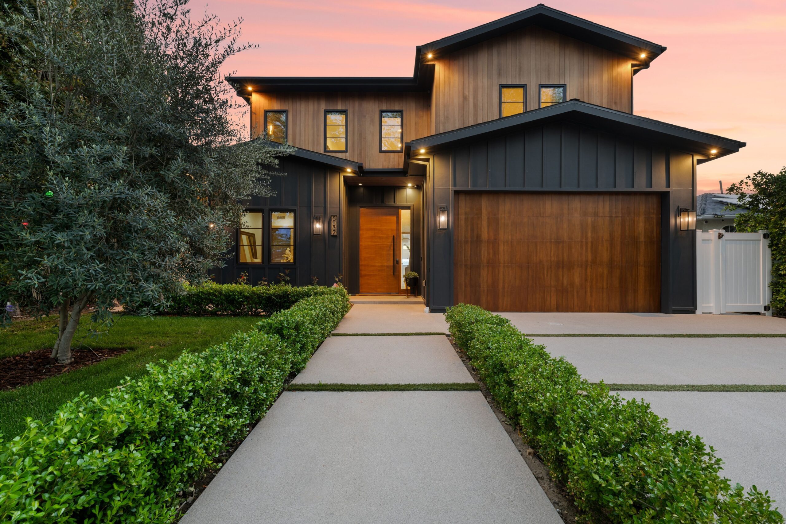 Modern two-story house with wood and dark panel exterior, large windows, and attached garage, set against a sunset sky with neatly trimmed hedges and a concrete driveway.